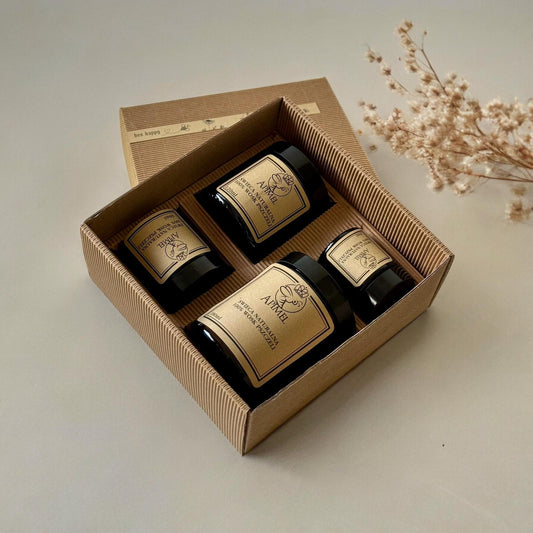 Zestaw czterech naturalnych świec w ciemnych szklanych słoikach z etykietą marki Apimel, umieszczony w estetycznym, ekologicznie wyglądającym pudełku z tektury falistej. Świece są starannie ułożone na beżowym tle obok gałązki suszonych kwiatów, co tworzy elegancką i minimalistyczną kompozycję. Zdjęcie zrobione z góry pudełka . Pudełko jest otwarte